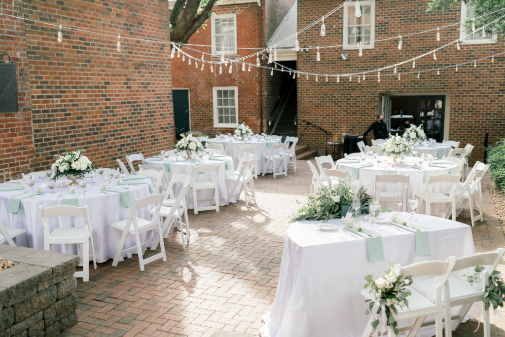 Round reception tables covered in white table clothes with sage green napkins are placed in the Linden Row Inn courtyard. White string lights hang above the tables. 