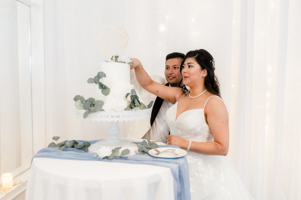 Veronica and Joshua are cutting their 1-tier wedding cake together. The cake is decorated with eucalyptus leaves and is on a round table with a blue runner. 