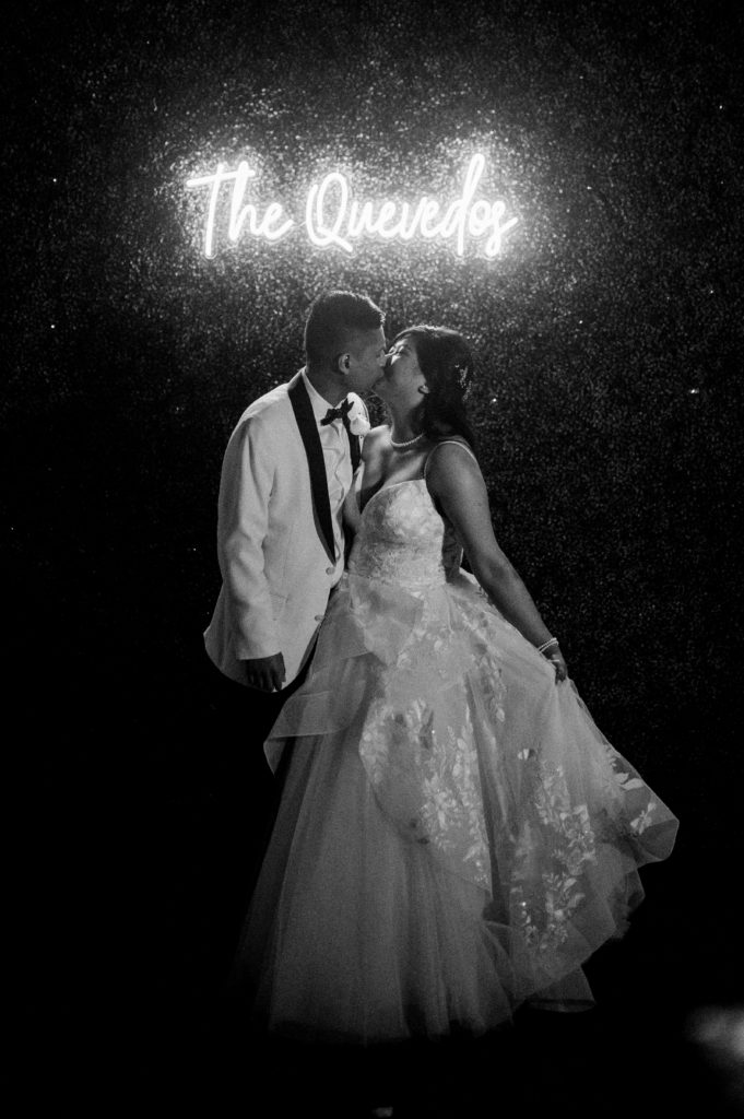 Joshua and Veronica are sharing a kiss at their rustic wedding under a neon sign that reads "The Quevedos." 