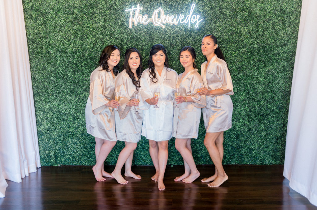 Veronica and her four bridesmaids pose in front of a greenery wall with a neon sign that reads, "The Quevedos." 