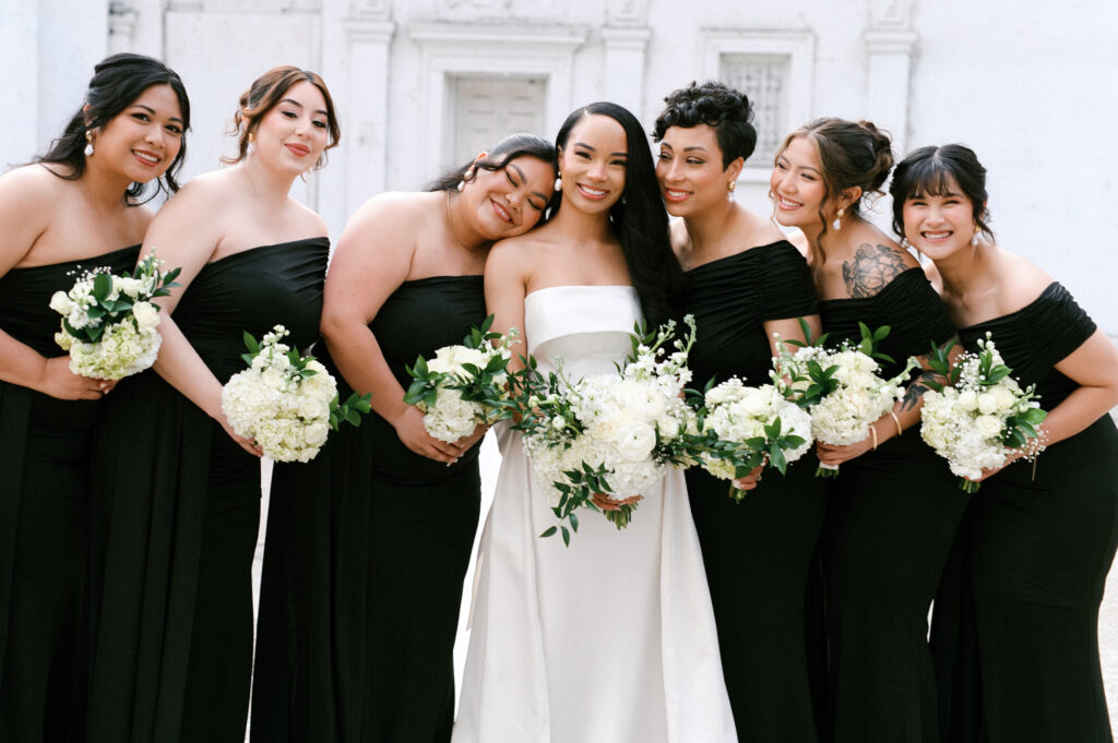 Bride surrounded by her bridesmaids in black dresses holding white bouquets at Gabriela and David’s Common House Richmond wedding by Chelsea Schmidt Photography.