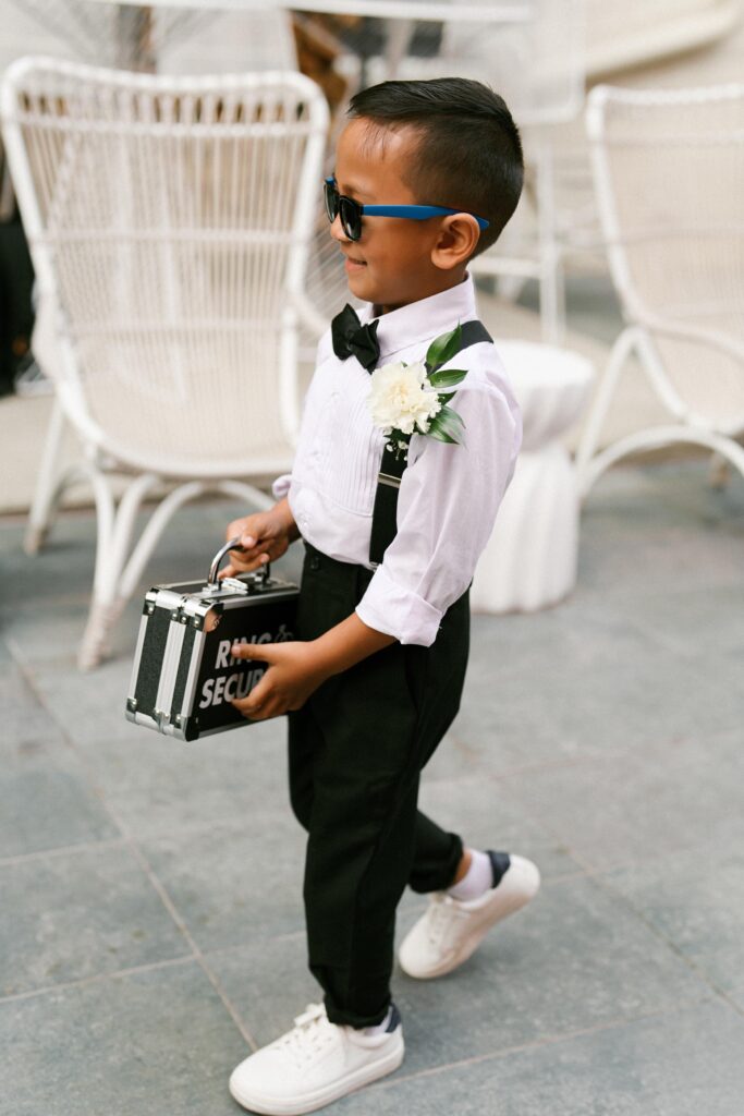 Ring security boy wearing sunglasses and carrying a mini briefcase while walking down the aisle at Common House Richmond.